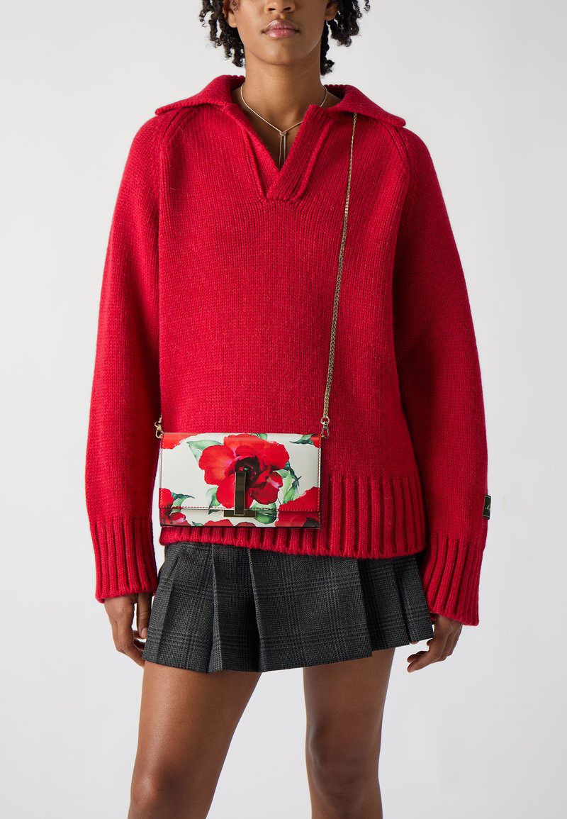 Maglione rosso lavorato a maglia con colletto, maniche ampie e orlo a coste; abbinato a una borsa a tracolla con motivo floreale e una gonna a pieghe scozzese.