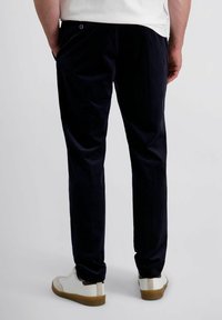 Man in een marineblauwe broek en witte sneakers, staand met zijn handen in de zakken tegen een plain lichte achtergrond.