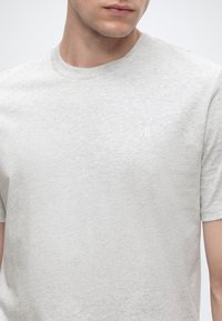 Joven con una camiseta gris claro de cuello redondo y un pequeño logo blanco "M OP" en el pecho izquierdo.