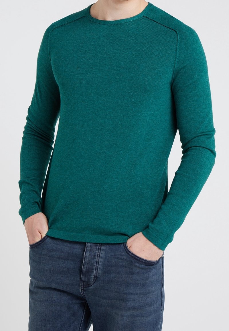 En man i en teal långärmad tröja och mörkblå jeans med händerna i framfickorna mot en enfärgad bakgrund.