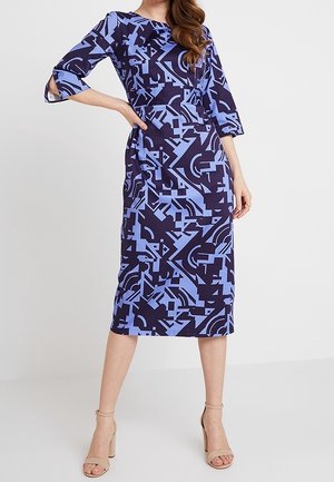 Vrouw draagt een knielange jurk met blauw-zwart geometrisch patroon, driekwart mouwen en beige sandalen met hoge hakken.