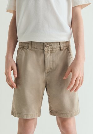 Persona con pantalones cortos beige hasta la rodilla y camiseta blanca, de pie con los brazos relajados a los lados contra un fondo neutro.