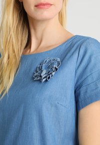 Sininen denimpaita, jossa on pyöreä pääntie, lyhyet hihat ja kerroksellisesta kankaasta valmistettu teksturoitu kukkaesine. Siinä on rintatasku.