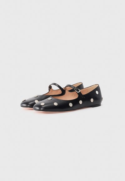 Black leather flats with a rounded toe and a Mary Jane strap, featuring large white polka dots and a flat sole. Smooth texture, minimalist design.