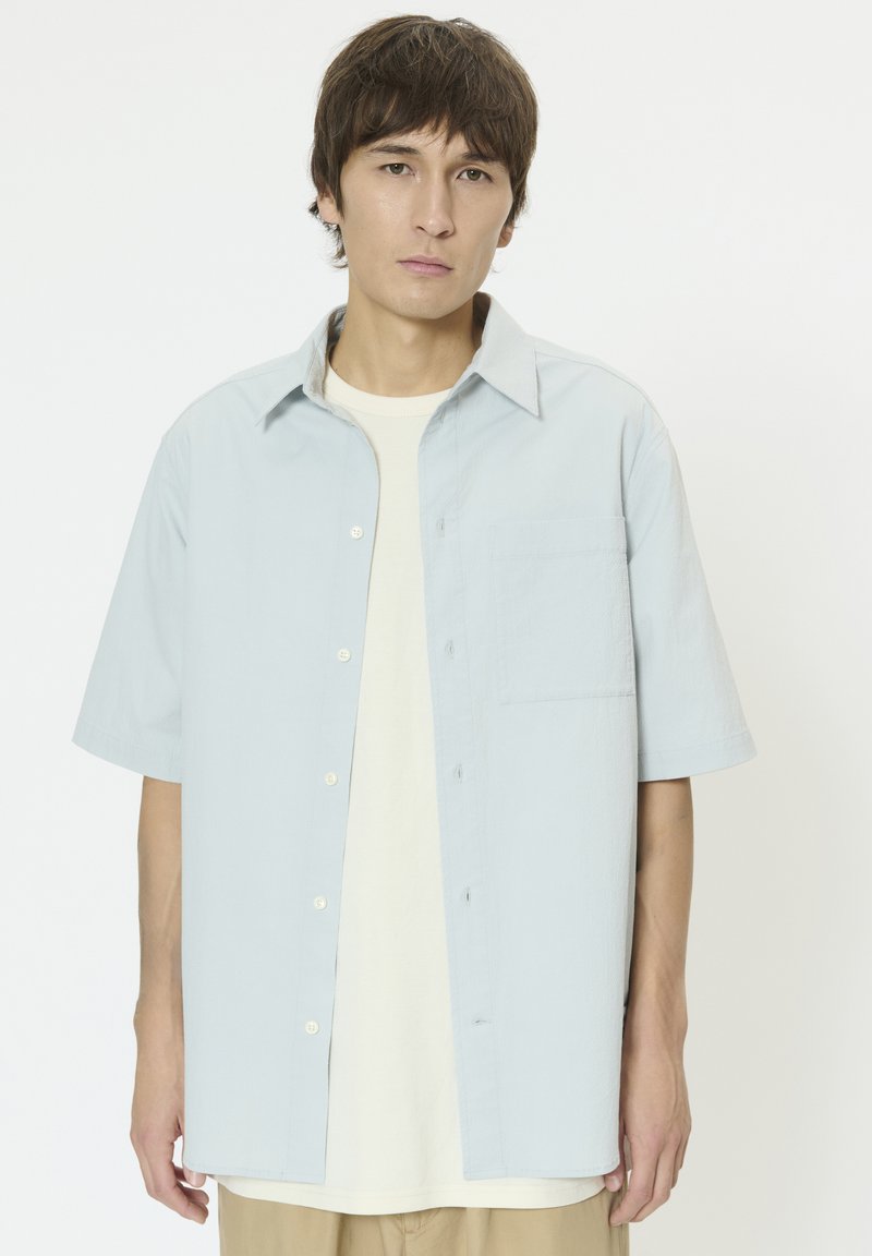 Hellblaues kurzärmliges Hemd mit Knopfleiste und einer Brusttasche, gefertigt aus strukturiertem Stoff. Wird über einem cremefarbenen T-Shirt getragen.