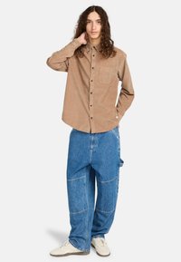 Braunes Cordhemd mit Knopfleiste und einer einzelnen Tasche, kombiniert mit übergroßen blauen Denim-Jeans mit Patchwork-Design.