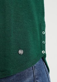 Grüner Strickpullover mit geripptem Halsausschnitt und seitlichen Knöpfen. Verfügt über ein rundes Etikett mit der Aufschrift "Esprit" auf der unteren linken Seite. Strukturierter Stoff.