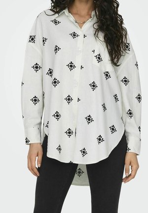 Femme portant une chemise blanche ample avec des motifs géométriques noirs brodés et un pantalon noir, se tenant debout avec les mains le long du corps.