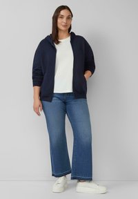 Marine blauwe zip-up hoodie over een wit T-shirt, gecombineerd met lichtblauwe flared jeans en witte schoenen met een gestructureerde zool.