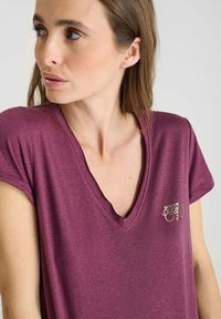 Vrouw met schouderlang bruin haar, gekleed in een paarse V-hals T-shirt met een klein geborduurd logo op de borst, kijkend naar links.