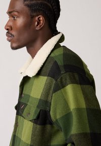 Chaqueta a cuadros verdes con un patrón de cuadros negro y oliva, que presenta un collar de felpa blanco y un bolsillo frontal. Confeccionada con una tela texturizada.