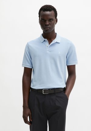 Jeune homme portant un polo bleu clair rentré dans un pantalon noir avec une ceinture noire, debout devant un fond blanc uni.