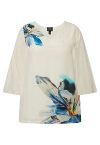 Hvid top med 3/4 ærmer, der har et blåt blomsterprint på den ene skulder. Lavet af letvægtsstof med en blød tekstur.