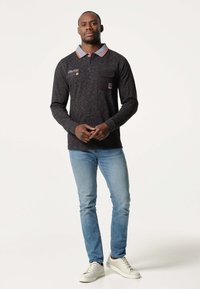 Polo noir à manches longues avec des accents de col rouges et gris, doté d'une poche frontale. Porté avec un jean bleu clair et des baskets blanches.