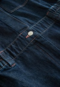 Gros plan sur un tissu en denim bleu foncé montrant un bouton en métal, des surpiqûres orange, des passants de ceinture et des coutures de poche sur un jean.