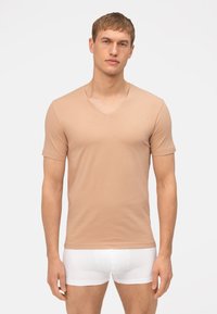 T-shirt beige a maniche corte con scollatura a V in tessuto morbido, caratterizzato da un design aderente. Modello abbinato a boxer bianchi, in piedi contro uno sfondo neutro.