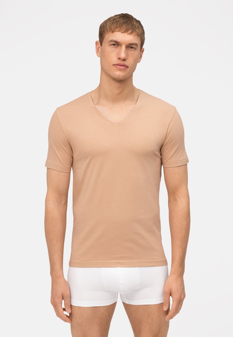 T-shirt beige a maniche corte con scollatura a V in tessuto morbido, caratterizzato da un design aderente. Modello abbinato a boxer bianchi, in piedi contro uno sfondo neutro.