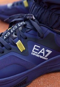 Sapatilha de desporto azul-marinho com parte superior sintética texturizada, design bicolor, destaque em amarelo e logótipo EA7 na lateral com atacadores em contraste.