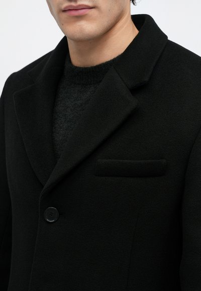 Άνδρας που φορά μαύρο μαλλί παλτό με τσέπη notch πάνω από ένα σκούρο πλεκτό πουλόβερ, με κουμπωμένο ένα μόνο μαύρο κουμπί ορατό.