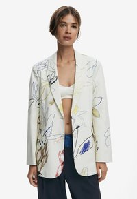 Blazer blanc avec des motifs floraux colorés en bleu, jaune et marron. Il présente une coupe décontractée, des revers en notch et deux poches avant.