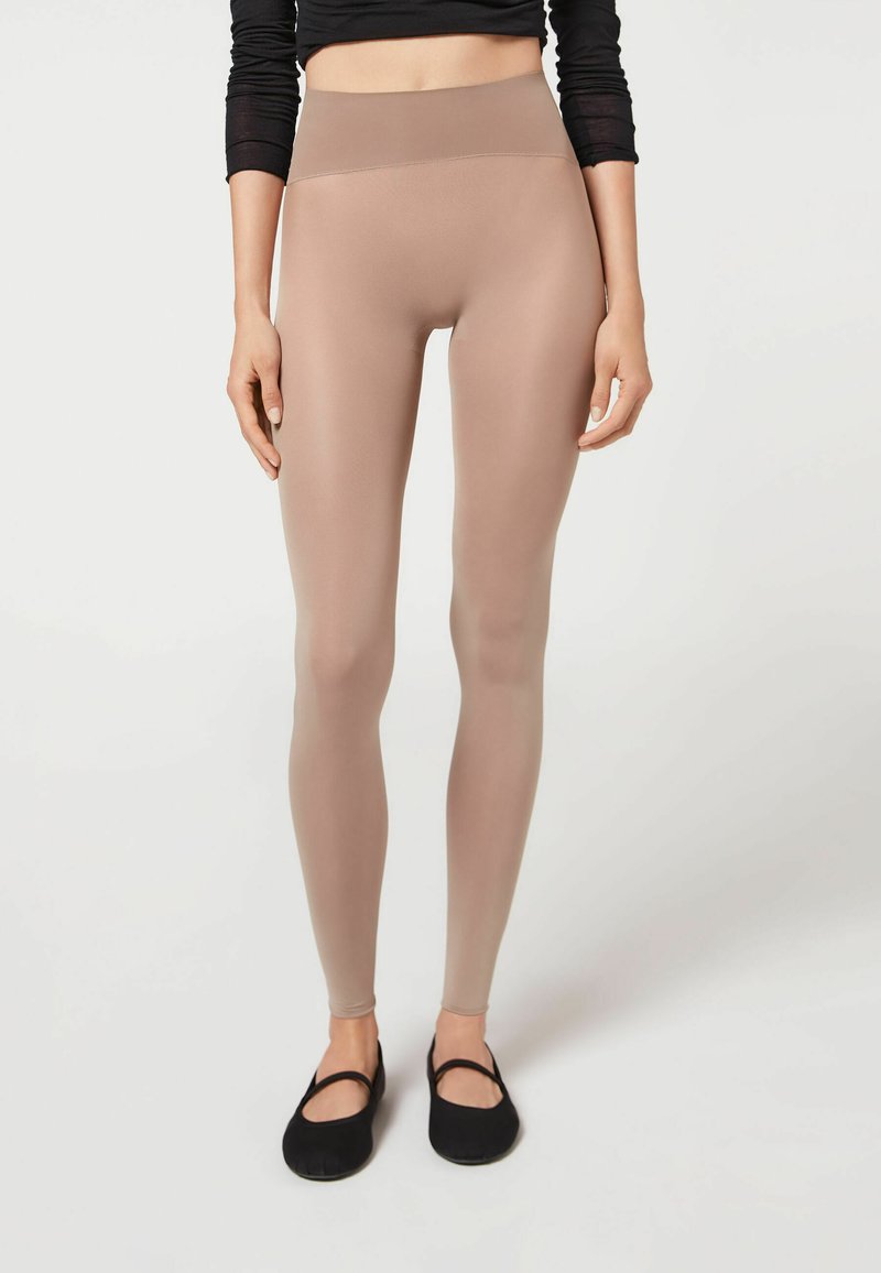 Beige højmidte leggins lavet af glat, strækbart stof, med et enkelt design og en kropsnær pasform, parret med sorte flade sko.