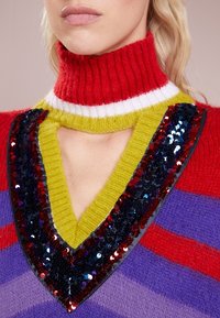 Dzianinowy sweter z czerwonym, prążkowanym golfem i żywymi paskami. Posiada dekolt w kształcie litery V ozdobiony zielonymi i cekinowymi detalami.