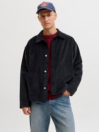 Black corduroy jacket with a collared neck, featuring silver snap buttons, front pockets, worn over a red knit sweater and blue jeans.