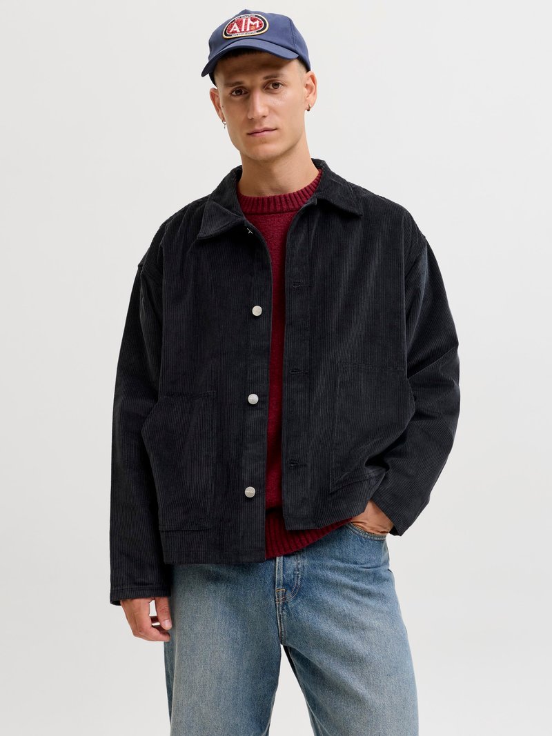 Black corduroy jacket with a collared neck, featuring silver snap buttons, front pockets, worn over a red knit sweater and blue jeans.