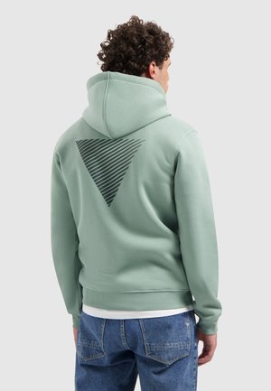 Man met krullend haar, gekleed in een lichtgroene hoodie met een zwart diagonaal gestreepte driehoek op de achterkant, gecombineerd met een blauwe spijkerbroek.