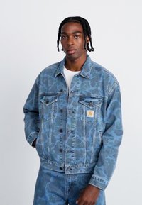 Blaue Jeansjacke mit strukturiertem Muster, ausgestattet mit Knöpfen, Brusttaschen und einem Logo-Patch. Getragen mit einem weißen Unterhemd.