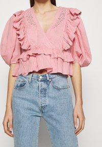 Růžová bavlněná blůza s volánovými rukávy a hlubokým výstřihem do V, zdobená krajkou a peplum spodním lemem, v kombinaci se světlými modrými džíny.