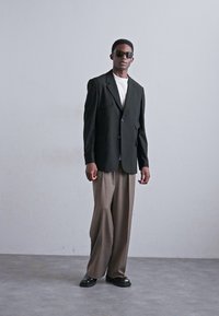 Black tailored blazer over a white t-shirt, paired with brown wide-leg trousers and black shoes. Model wears dark sunglasses.