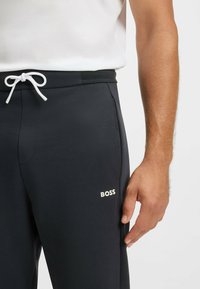 Svarta träningsbyxor i mjukt material, med en vit dragkedja och en "BOSS"-logotyp i krämfärg på vänstra låret.