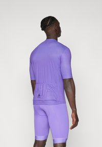 Kortsleevet lilla treningstrøye med baklomme. Glatt tekstur, kroppsnær design, og merket logo nederst på ryggen.