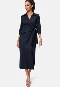 Navy wrap dress made of smooth fabric featuring a collar, three-quarter sleeves, a tied waist, and a side slit.