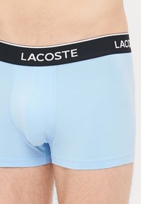 Svetlo modri boksarji narejeni iz mehkega materiala z črnim elastičnim pasom, ki nosi belo napisano „LACOSTE“.