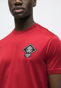 Rød t-shirt med korte ærmer i bomuldsblanding med rund halsudskæring. Har en sort diamantformet lap med hvidt broderet logo.
