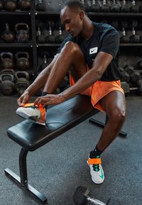 Atleta sentado en un banco negro, vistiendo zapatillas multicolores con una banda naranja prominente, una camiseta negra y pantalones cortos naranjas.