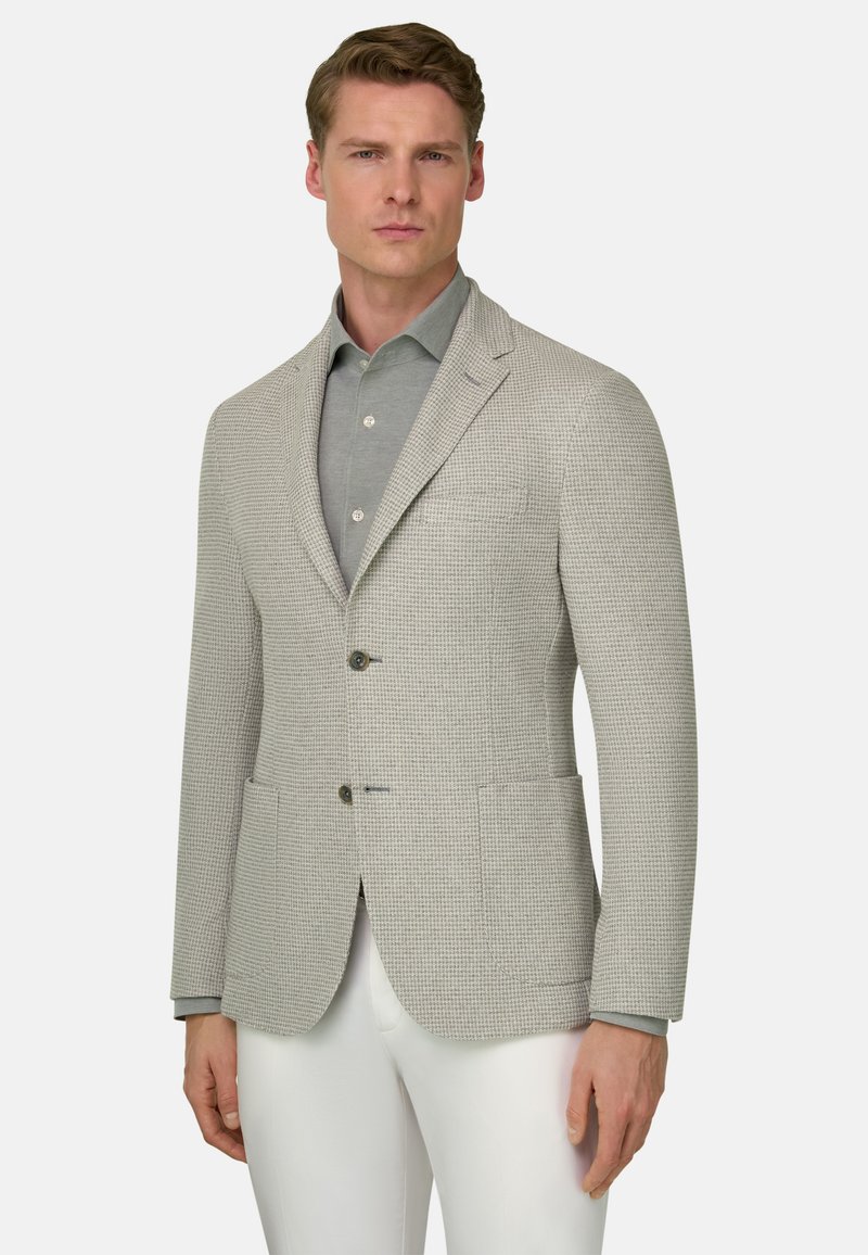 Man draagt een beige textuurblazer over een grijs overhemd met knopen en witte broek, staand tegen een effen lichte achtergrond.
