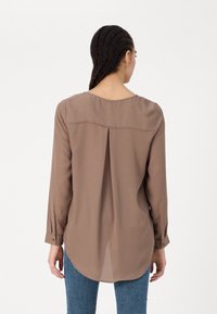 Femme avec des cheveux tressés portant un haut taupe à manches longues avec un pli dans le dos et un jean bleu, se tenant de dos contre un fond blanc.