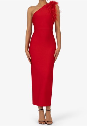 Vrouw draagt een nauwsluitende, asymmetrische rode jurk met veerdetail op de schouder en zwarte enkelbandhakken.