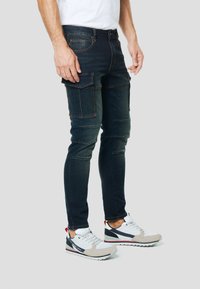 Tmavomodré cargo džínsy s viacerými bočnými vreckami, úzky strih a kontrastné prešitie. Doplněné bielymi a sivými športovými teniskami.