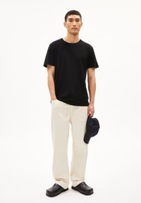 Schwarzes kurzärmliges T-Shirt, cremefarbene Jogginghose mit zwei Vordertaschen, schwarze Clogs zum Hineinschlüpfen und eine marineblaue Baseballmütze in der Hand gehalten.