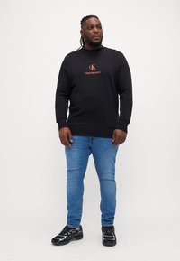 Crna dukserica s logom Calvin Klein Jeans, plave uske traperice i crne atletske cipele s ljubičastim akcentima. Ležerna odjeća prikazana na modelu.