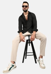 Camicia a quadri nera, pantaloni beige e sneakers eleganti con dettagli verdi. Modello seduto su uno sgabello nero, rivolto in avanti.