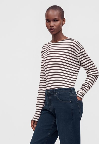 Femme aux cheveux courts portant une chemise à manches longues blanche et marron rayée et un jean bleu foncé, debout avec une main dans la poche.