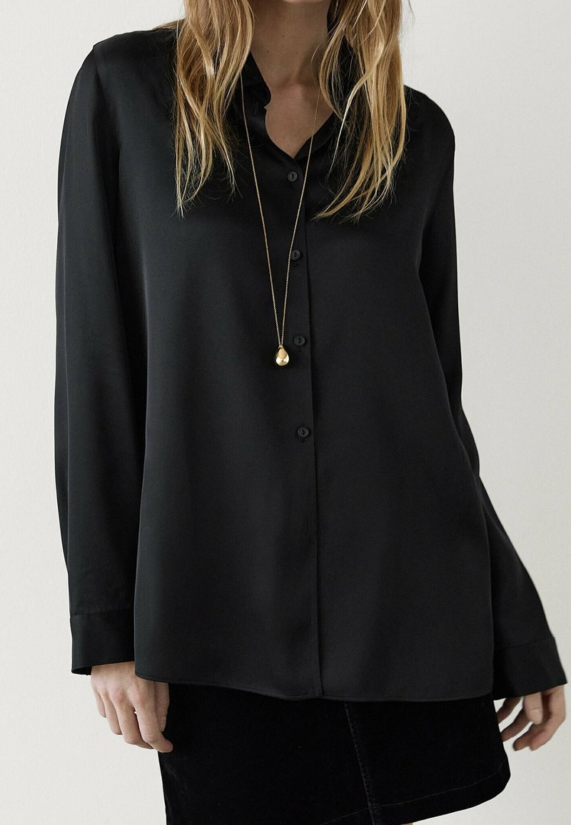 Femme portant un chemisier noir à manches longues boutonné, une jupe noire et un long collier en or avec un pendentif en forme de goutte.
