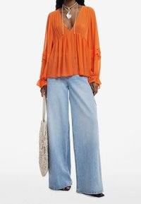 Blusa in chiffon arancione con scollatura a V, maniche lunghe e dettagli plissettati, abbinata a jeans in denim azzurro a gamba larga e una borsa intrecciata.