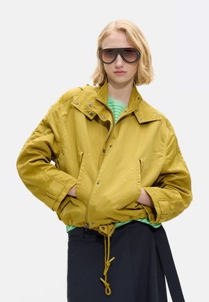 Osoba o blond włosach, nosząca duże czarne okulary przeciwsłoneczne, żółtą, oversize'ową kurtkę, zieloną koszulę w paski oraz ciemną spódnicę, z rękami w kieszeniach.