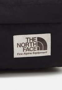 Sac en tissu noir avec une étiquette rectangulaire crème portant "THE NORTH FACE" et le logo de montagne en marron foncé. Surface texturée, bords cousus.